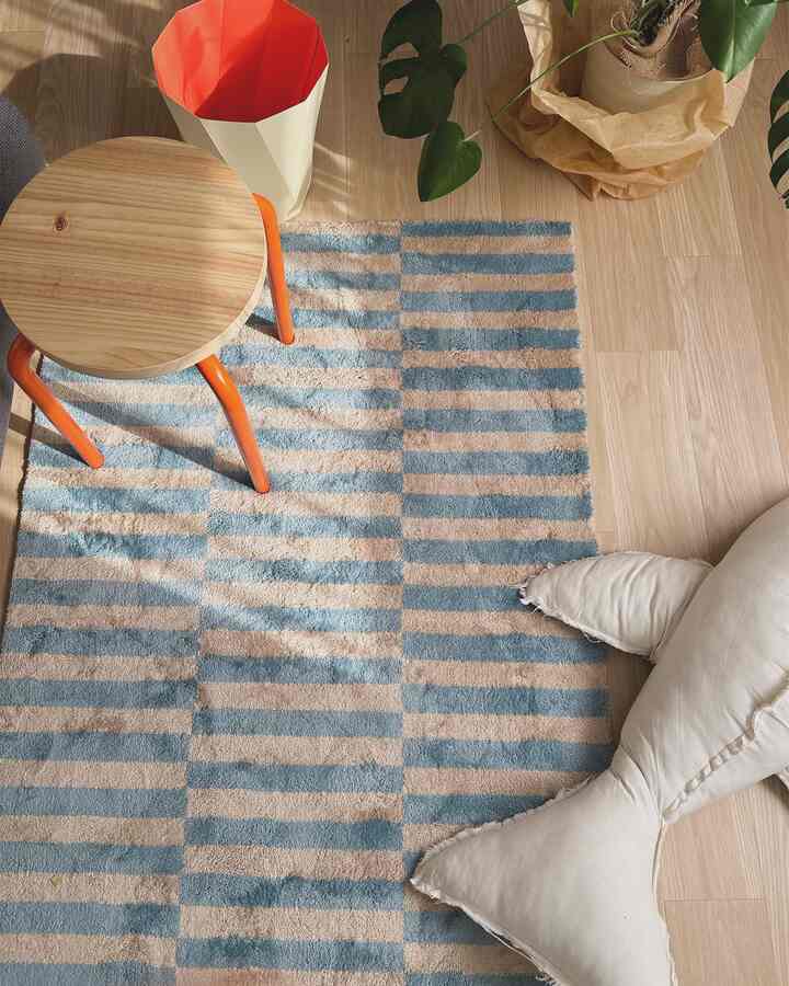 Pastel blue and beige striped rug on natural wood-tone floor, featuring orange-legged stool and plant, creating a cozy, natural interior