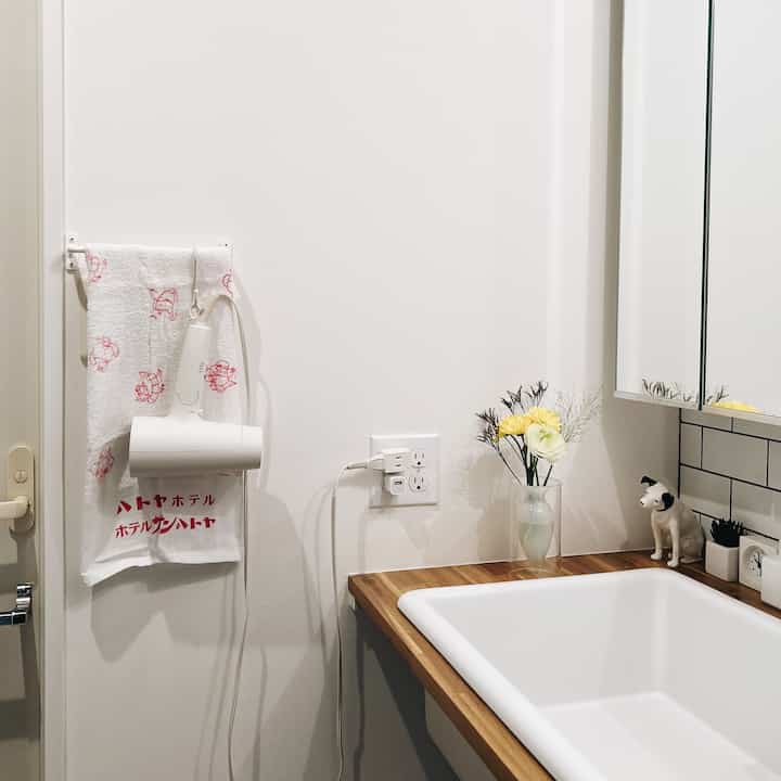 白とナチュラルウッドのシンプルなバスルーム、洗面台の上に花瓶とヘアドライヤーが置かれた清潔感のある空間