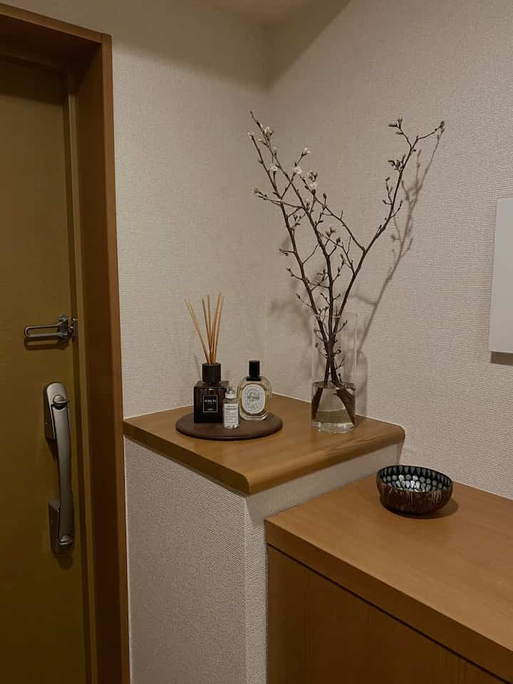 ベージュ色の壁とブラウンの木製家具が配置されたシンプルでナチュラルな玄関、透明なガラス花瓶とディフューザーが置かれている印象的な空間