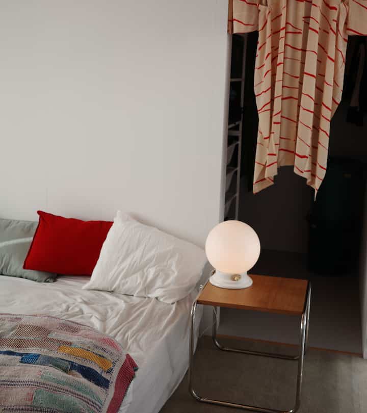 White-toned bedroom featuring a white bed with red cushion and a bedside table with spherical lamp in a minimal setting