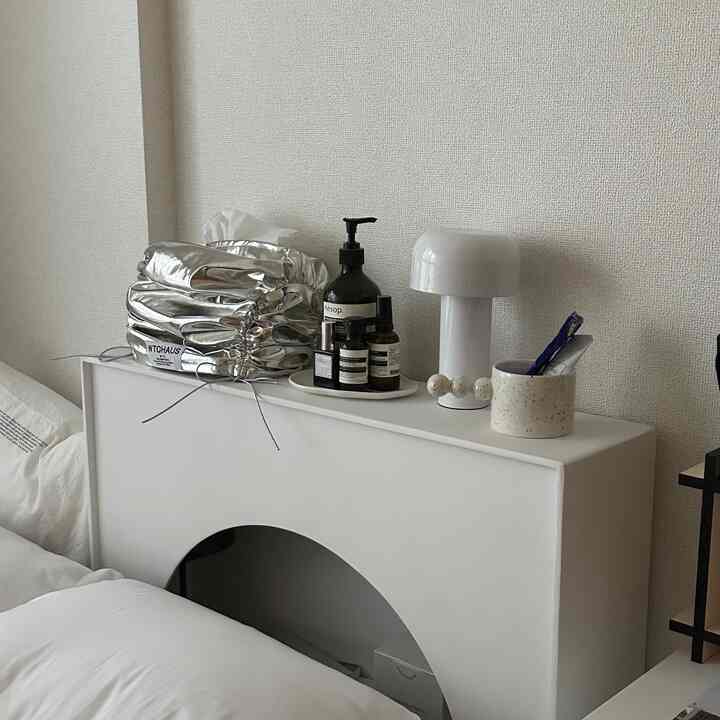 A bright white and natural toned bedroom featuring a minimalist bed and nightstand with a table lamp, creating a clean atmosphere