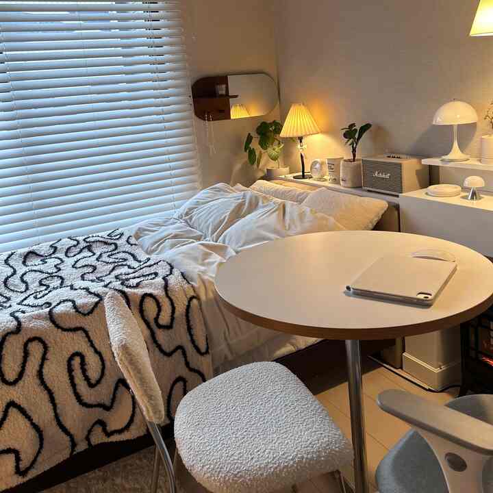 暖かいベージュ調の寝室、小さな丸テーブルとふわふわの白いスツール、ナイトテーブルと植物、ブラインドが配置された印象的な空間