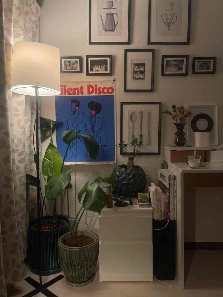 暖かい照明が灯る白い壁と木目調カーテンの落ち着いた空間、多様な額縁と観葉植物が配置されたホームオフィス兼くつろぎのインテリア