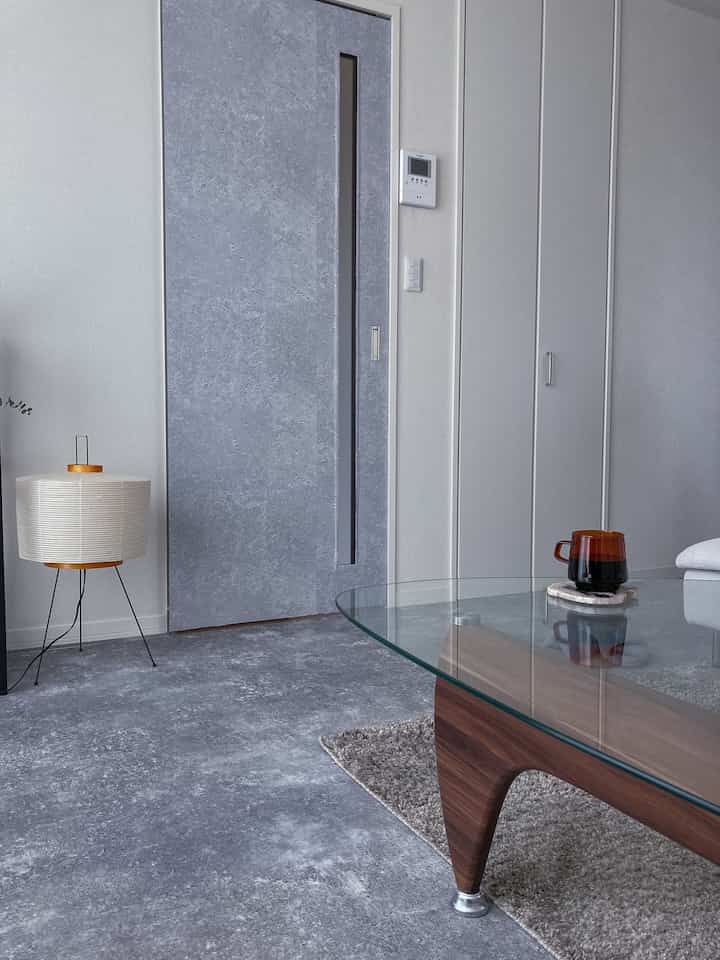 グレーのトーンが特徴的なリビング、ガラス天板のコーヒーテーブルと木製の脚、三脚のフロアランプが配置されたシンプルなインテリア。