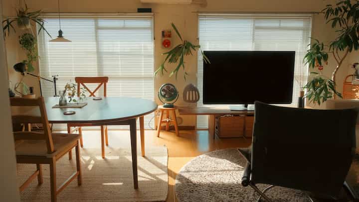 ナチュラルな色調の1LDKダイニングリビングルーム、木製家具とブラインド、植物、レトロなサーキュレーターが配置された雰囲気のある空間。