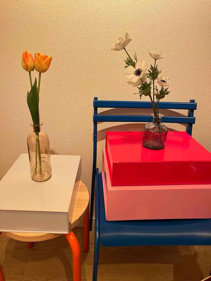 白い壁を背景に、木製スツールと青い金属製の椅子の上に花瓶とギフトボックスが置かれた温かみのある空間