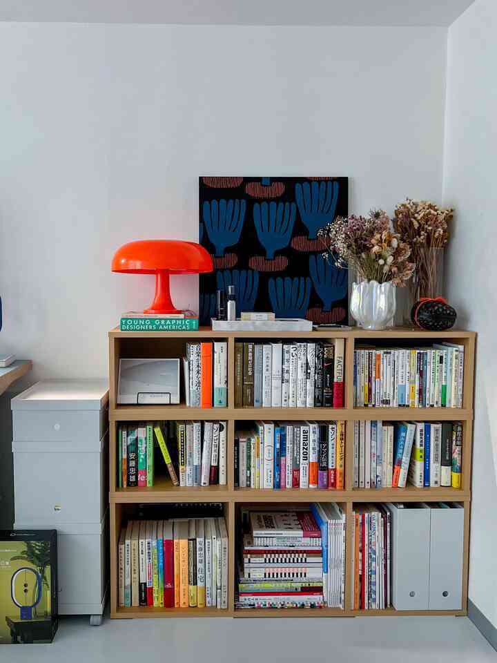 白い壁と木目調の本棚がある北欧風ワークスペース、オレンジ色のテーブルランプと花瓶がアクセントの整った空間