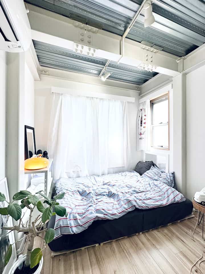白と青のトーンのワンルーム寝室、ストライプ柄の掛け布団カバーと植物が配置された清潔感と自然な雰囲気のあるインテリア