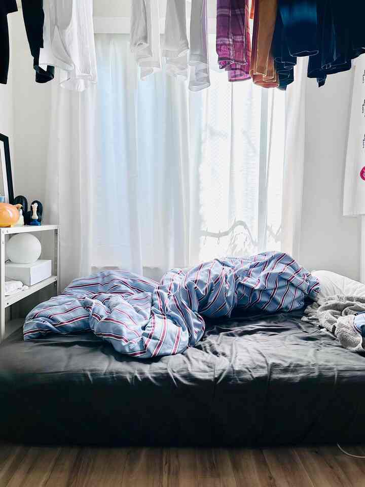 白と黒を基調とし、青のストライプ柄掛け布団カバーがある小さな寝室。窓際のカーテンと吊るされた洋服がある落ち着いた雰囲気の空間