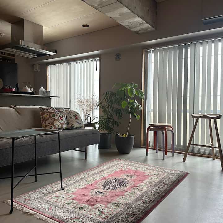 Natural-toned living room featuring gray sofa, pink rug, plants, and stools in a modern setting