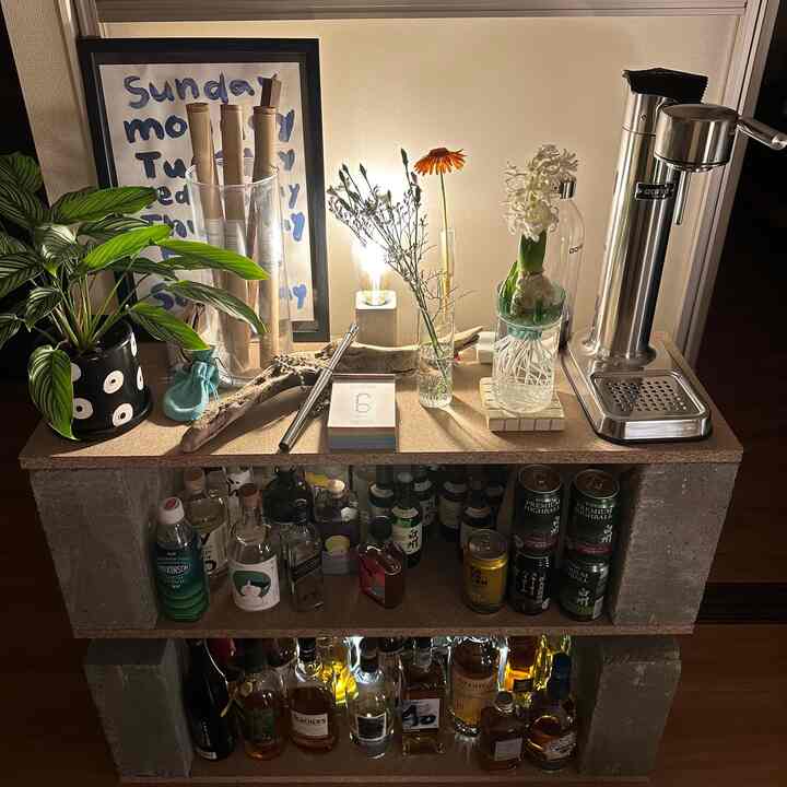 暖かい照明の自然なトーンが特徴の狭い部屋、ホームバーのディスプレイラックと植物、日めくりカレンダーが配置された雰囲気のあるインテリア