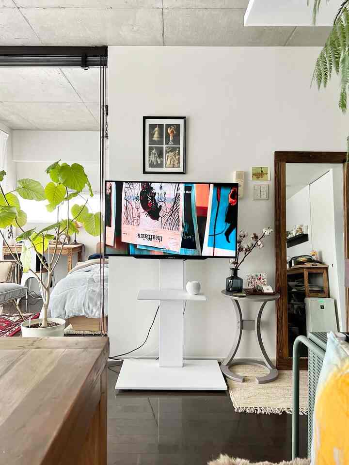 白と茶色を基調とした1人暮らしのリビング兼寝室、ミニマルなテレビ台とナチュラルな観葉植物が配置された落ち着いた印象の空間