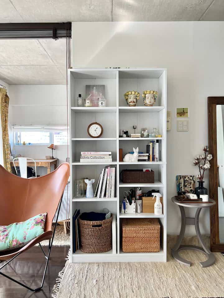 A modern studio living room with white and brown tones featuring a bookshelf, brown leather butterfly chair, and a side table in an organized setting