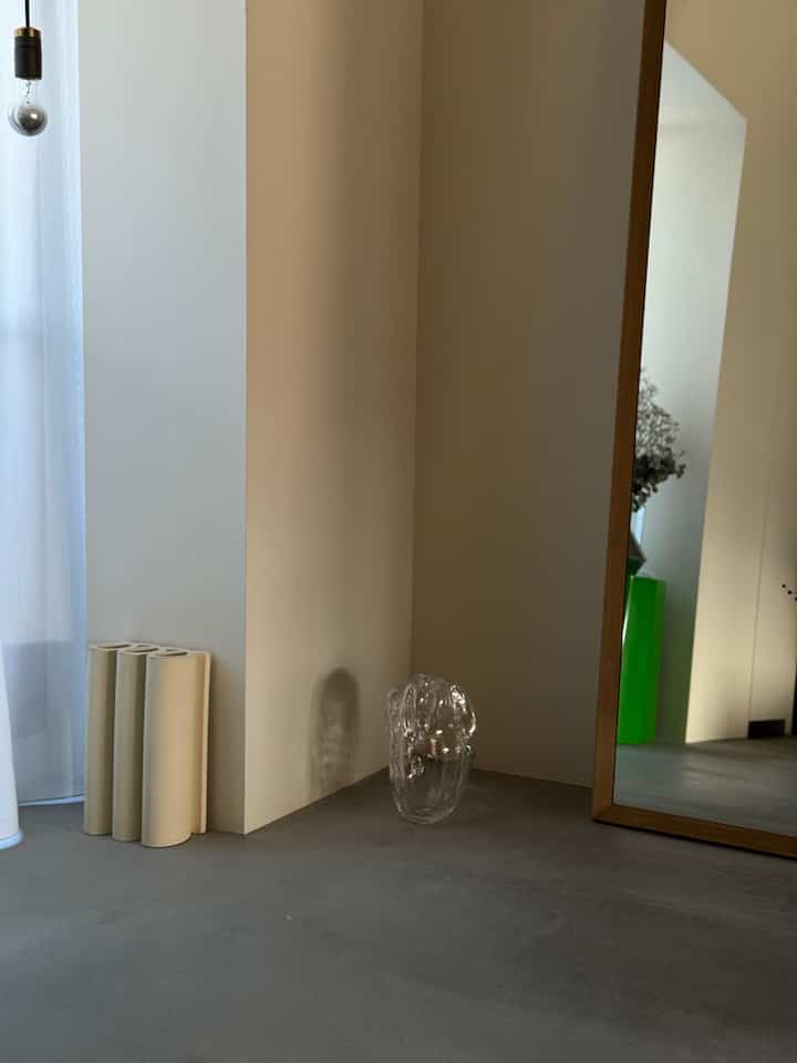 ベージュの壁とグレーのモルタル床の玄関、木製フレームの鏡と透明なガラス花瓶が配置されたシンプルでモダンなインテリア