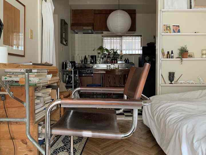 Natural-toned studio combining bedroom and kitchen, featuring a brown leather chair and white bedding in a cozy space