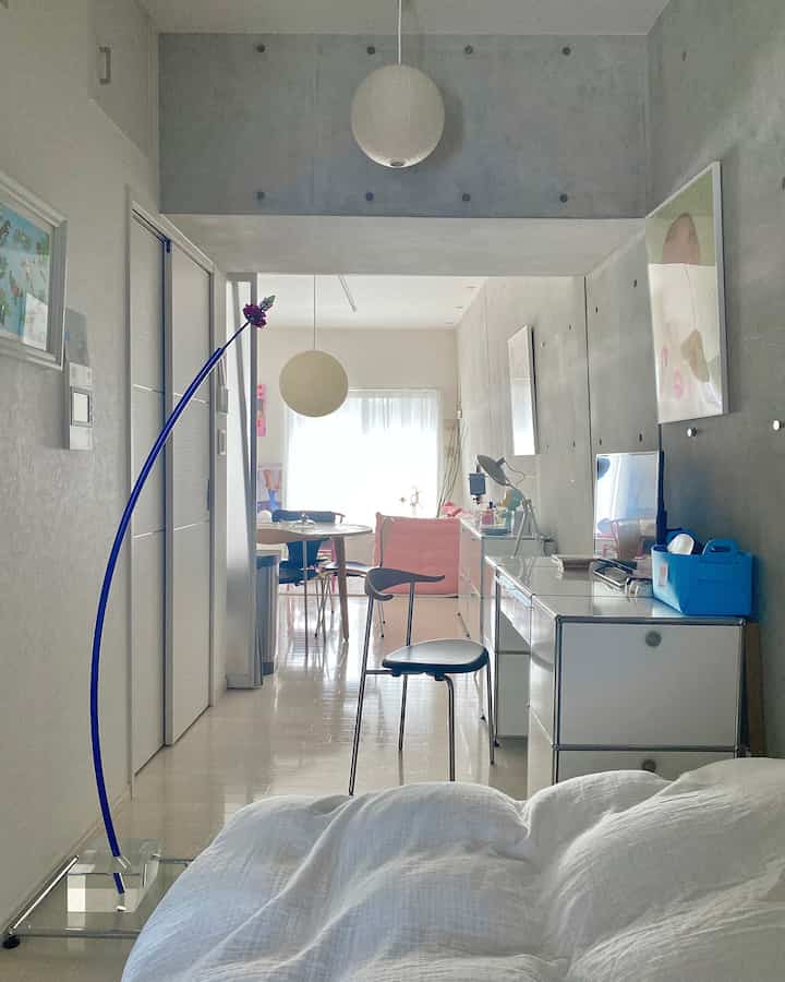 グレーの無機質な壁と白い寝具があるワンルームのワークスペース、ペンダントライトとモダンな家具が配置された明るく清潔な空間