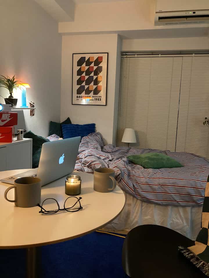 白い壁と青いカーペットが調和するワンルームの寝室、ベッドとテーブル上のノートパソコンとキャンドルが落ち着いた雰囲気を醸し出す空間