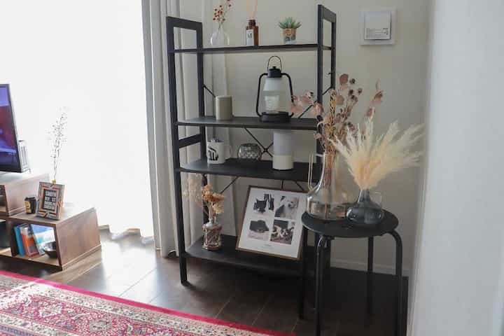 白い壁と茶色い床のリビングに、黒いメタルラックとスツール、自然素材の飾りが配置された雰囲気のあるインテリア