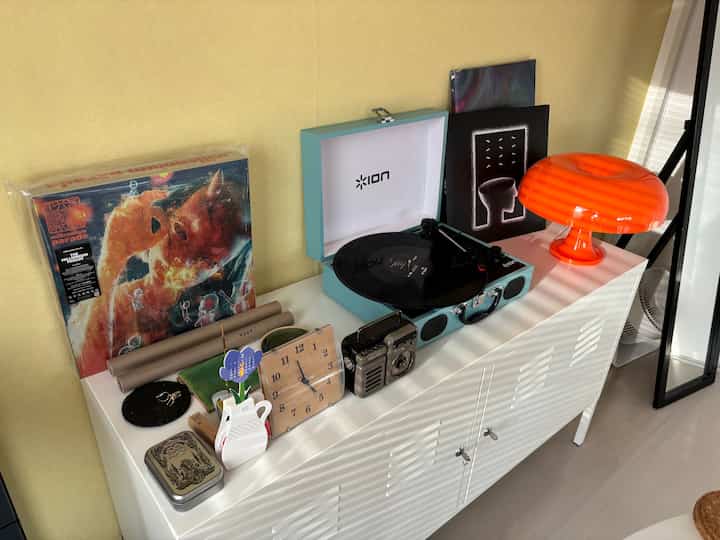 黄色い壁と白いキャビネットがあるリビング、ターンテーブルやヴィンテージ時計、オレンジのランプが配置されたレトロな雰囲気のインテリア