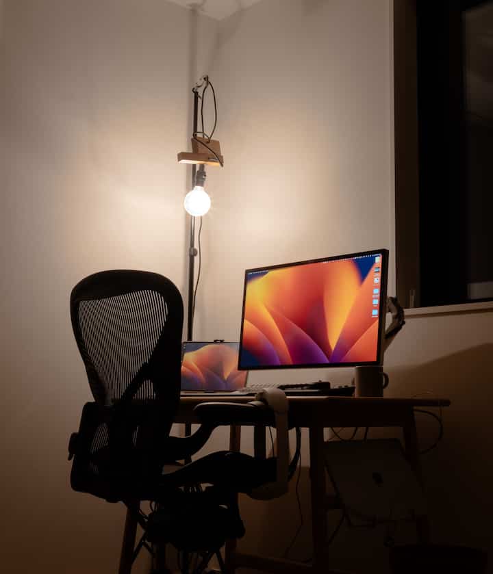 白い壁と木目調の机があるモダンなホームオフィス、エアロンチェアとAppleスタジオディスプレイが配置された集中できる空間