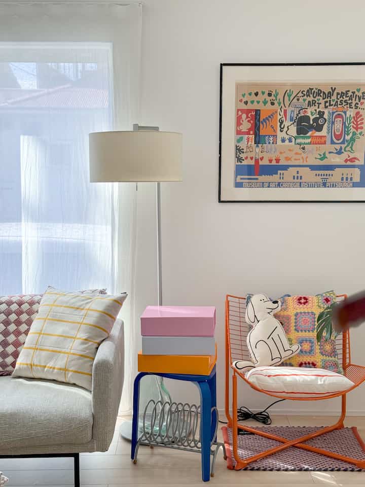 白とオレンジ、青の色調のリビング、ソファとスツール、アームチェア、カラフルなクッションと絵画が配置された雰囲気あるインテリア