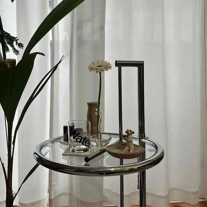白いカーテンを背景に、花と小さなディフューザー、植物が置かれたクロームとガラスの丸いサイドテーブルのあるシンプルなリビングの一角