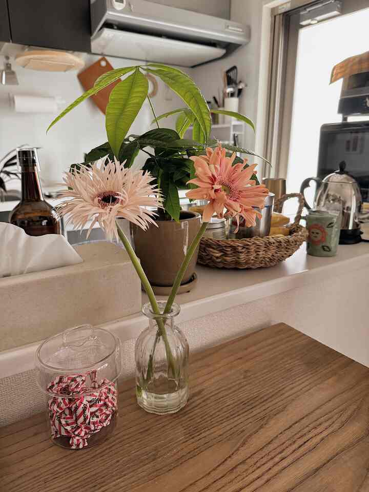 ナチュラルな色調のキッチン、透明なガラスの花瓶にピンクのガーベラと緑の植物が配置された雰囲気のあるインテリア