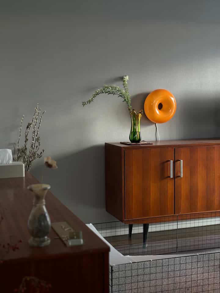 Cozy mid-century modern living room with gray walls, wood-tone console table, orange wall lamp, and green vase with flowers