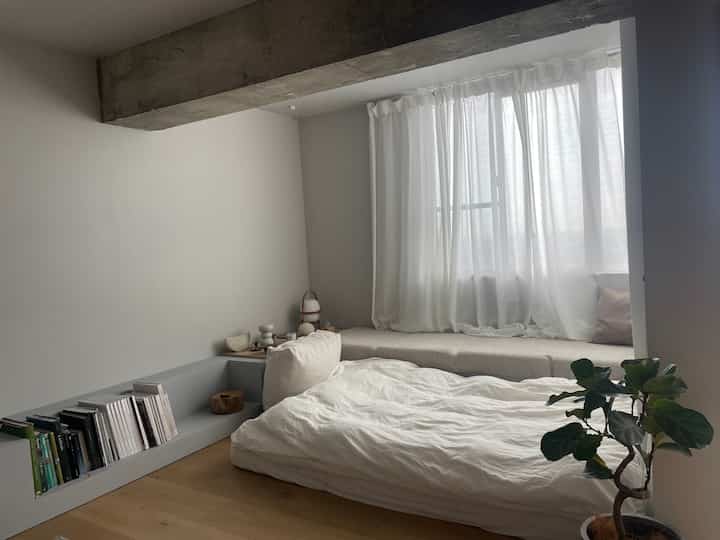 Natural modern living room in white and beige tones featuring a window daybed with cushions, bookshelf, and potted plant