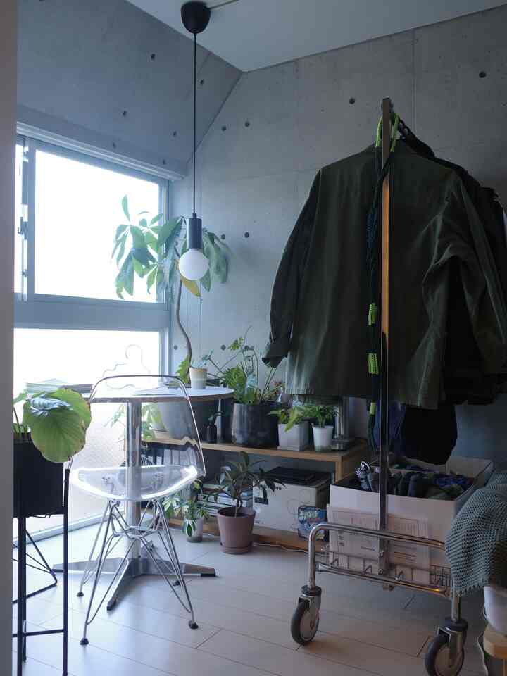 白い床とコンクリート壁が際立つコンパクトな部屋、植物と透明な椅子、コートラックが置かれたシンプルなホームオフィス空間