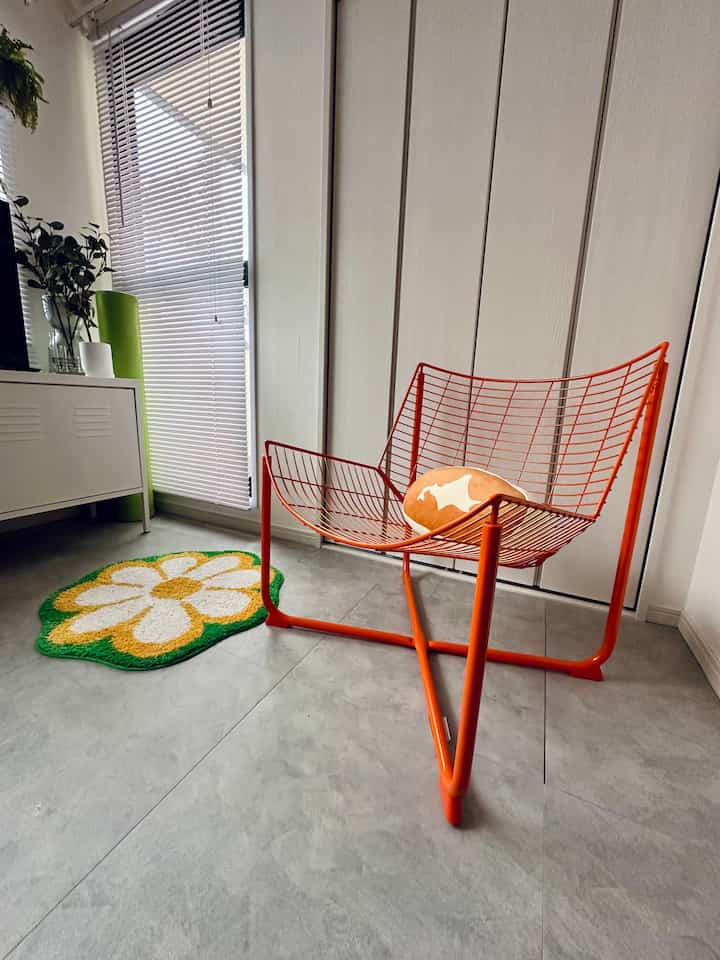 明るいオレンジ色の金属製アームチェアと白いブラインド、花柄のラグが配置された清潔感のある玄関スペース