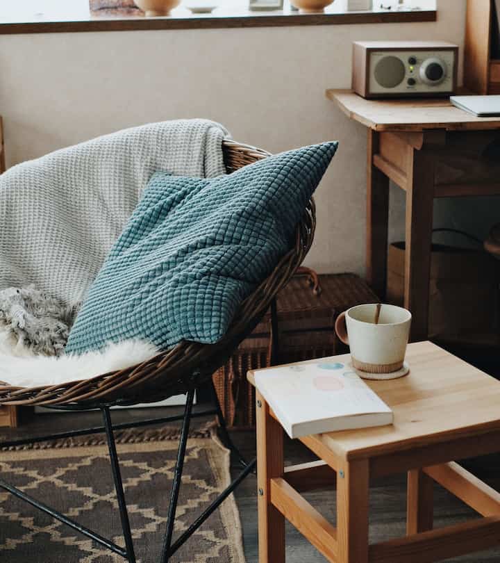 Beige and brown toned living room corner featuring a rattan armchair with cushions and a wooden stool holding a mug and book, cozy reading spot