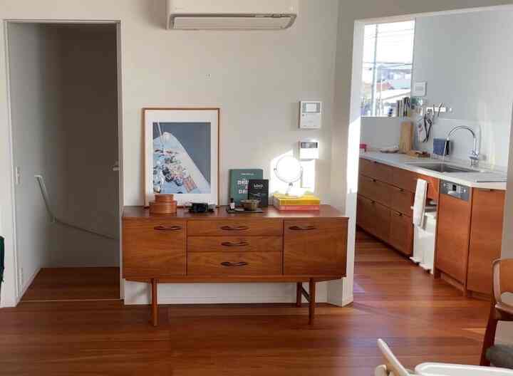 白い壁と木目調の床、キッチンとリビングが繋がるナチュラルな空間、中央に木製の収納家具が配置されている