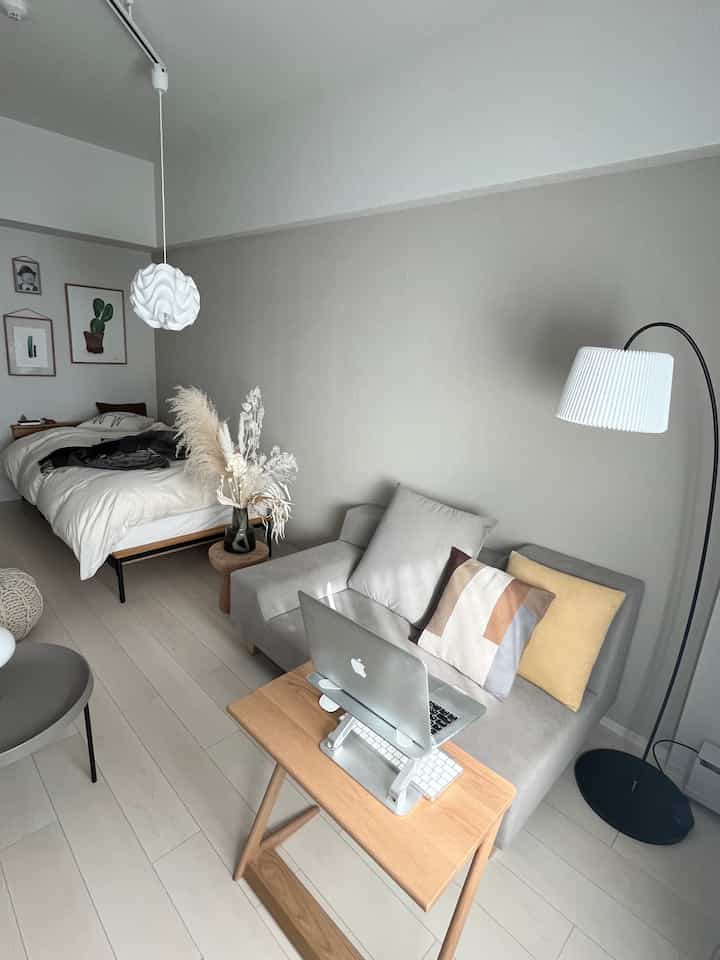 Light gray and wood-tone studio apartment featuring a bed, sofa, floor lamp, and laptop workspace in a minimalist setting