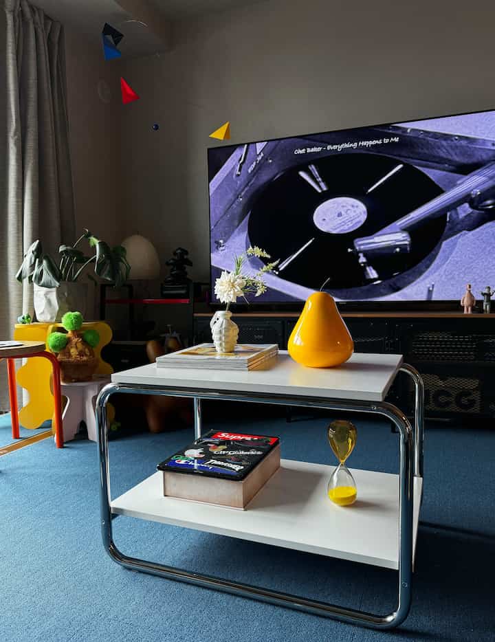 Blue carpeted living room featuring white side table, colorful mobile, and yellow decorative objet with modern atmosphere
