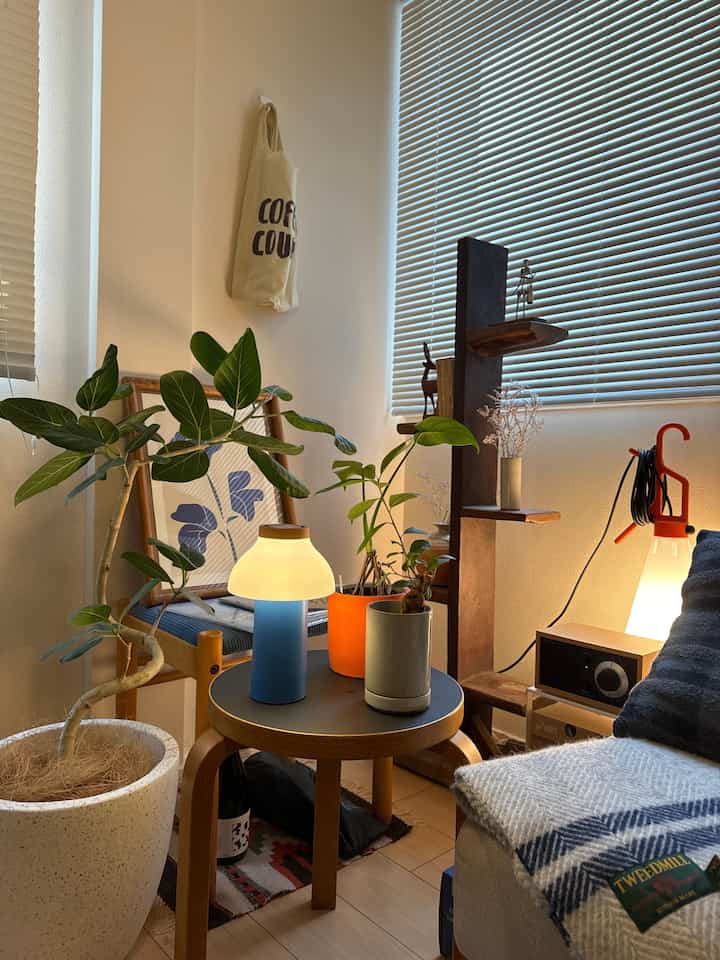 自然光が差し込むナチュラルトーンのリビング、中央に植物とブルーのテーブルランプが置かれたスツールがあり、温かみのある雰囲気のインテリア