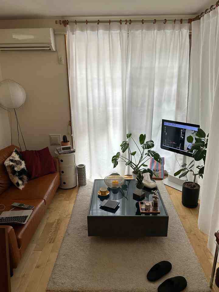 Mid-century modern living room with wide white curtains, brown leather sofa, black coffee table, and green plants arranged naturally