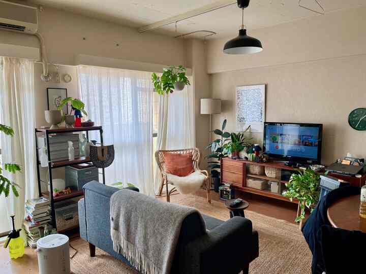 ベージュ色の壁とカーテンがあるワンルームのリビング、ブルーのソファとナチュラルな木製家具、植物が配置された自然な雰囲気のインテリア