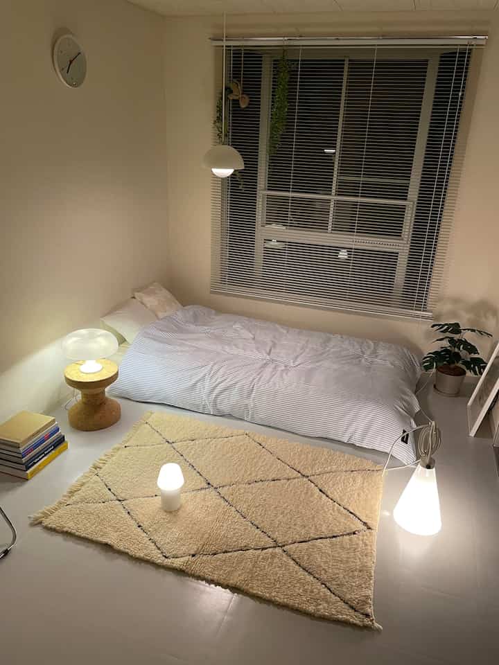 ナチュラルな色調の寝室、モノトーンの寝具と複数の照明が配置された落ち着いたミニマルな空間