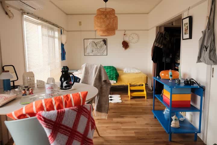 白と黄色のトーンのリビング、中央にベッドと青いオープンシェルフが配置された雰囲気のある空間