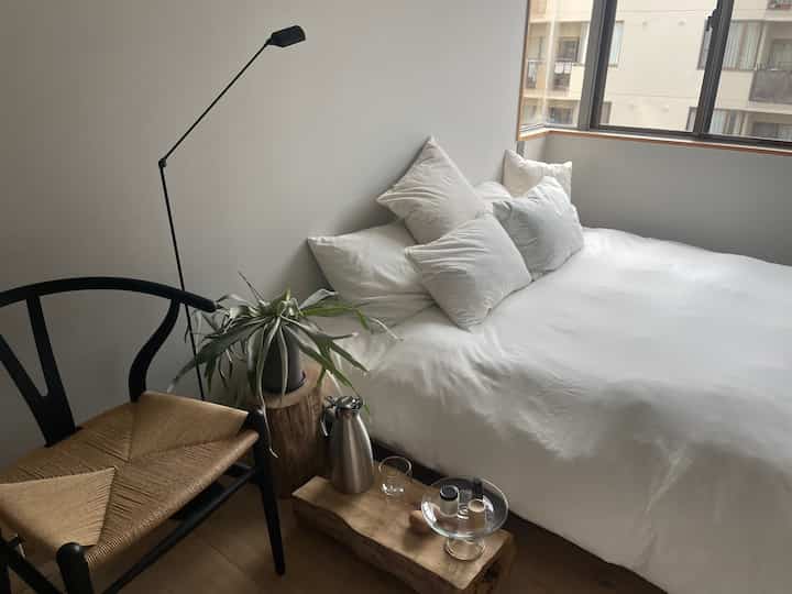 ホワイトとナチュラルトーンの寝室、Yチェアと木製スツールのサイドテーブル、モダンなフロアランプが配置された雰囲気のあるインテリア