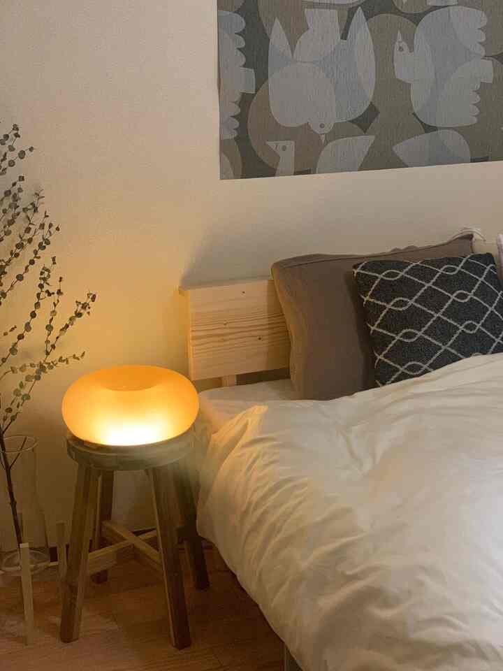 暖かいナチュラルカラーの寝室、木製スツールの上にLEDテーブルランプと白い寝具が配置された落ち着いた空間