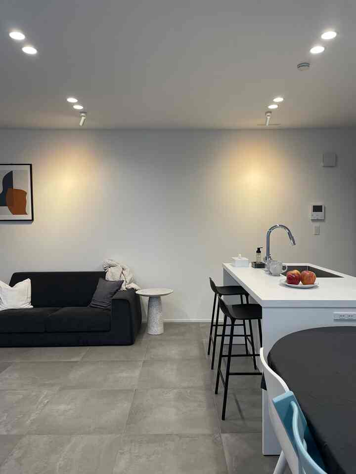 白い壁とグレーのタイル床のモダンなリビングとキッチン空間、黒いソファとバースツールが配されたシンプルなインテリア