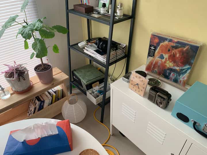 ベージュの壁と白い収納棚があるコンパクトな部屋、窓際には観葉植物と本棚が配置されたミニマルで清潔感のあるインテリア