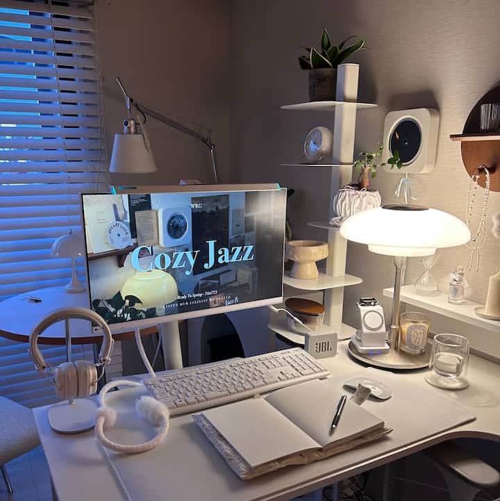 A white and beige toned home office space featuring a white desk, monitor, and table lamp creating a cozy workspace