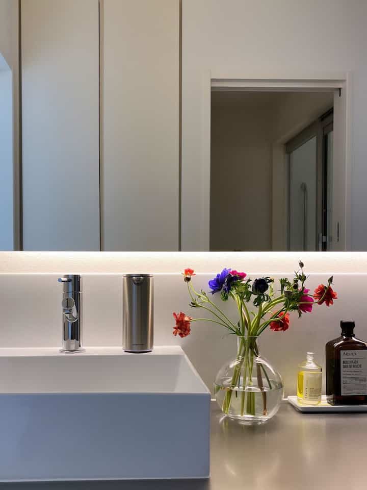 白とシルバーのトーンのバスルーム洗面スペースに透明な花瓶とカラフルな花が置かれた、清潔で洗練された雰囲気のインテリア