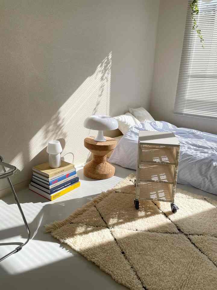 白とナチュラルカラーのコンパクトな寝室、床にマットレスとコルクのナイトテーブル、ベージュのラグが配置された明るい空間