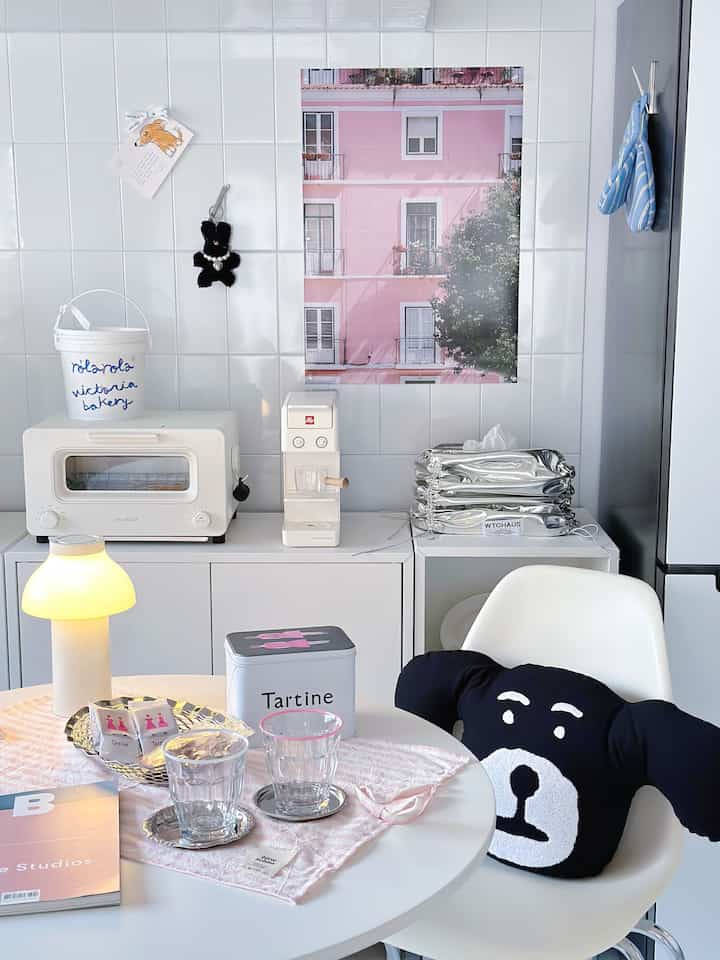 A modern kitchen space with white tiled walls featuring a pink poster, a white round dining table and a table lamp creating a cozy atmosphere.