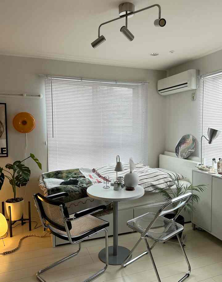 白とナチュラルカラーの調和が取れた小さなワンルーム寝室、透明な椅子と丸いテーブルが配置されたモダンな印象的な空間