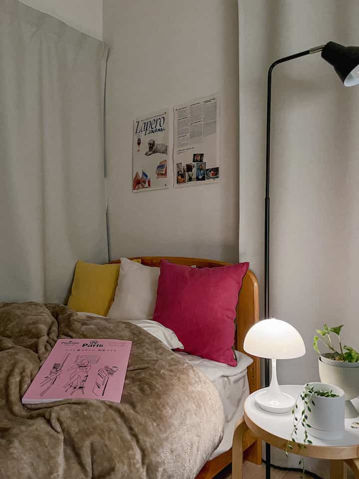 Cozy bedroom with white curtains, brown bedding, wooden bed frame, bright pink cushion and warm table lamp lighting